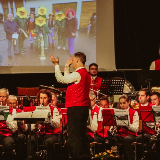 Ein Mann in einem roten Westchen steht vor einem Orchester und dirigiert, während ein Bild von Menschen in Kostümen auf einer Leinwand hinter ihm angezeigt wird.