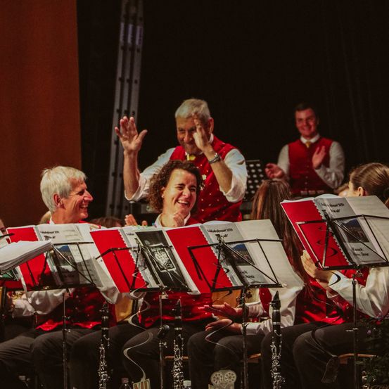Eine Gruppe von Musikern sitzt auf Stühlen, einige stehen und klatschen vor einem schwarzen Vorhang. Einige halten Notenblätter und andere halten Musikinstrumente wie Klarinetten.