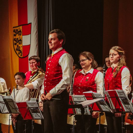 Ein Mann in Rot steht in der Mitte einer Gruppe von Kindern, sie halten Musikinstrumente und scheinen für ein Konzert zu proben.