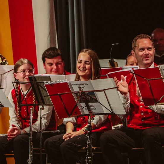 Eine Gruppe von Musikern in roten und weißen traditionellen Kostümen sitzt vor Notenständern, lächelt und schaut in die Kamera.