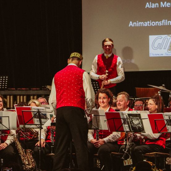 Eine Gruppe von Musikern, alle in roten Westen und schwarzen Hosen gekleidet, spielt ihre Instrumente auf der Bühne. Eine Projektionsleinwand mit Text im Hintergrund zeigt eine Präsentation.