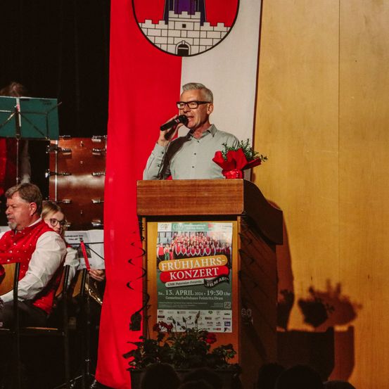 Ein Mann steht an einem Podium und spricht in ein Mikrofon. Dahinter ist eine rot-weiße Flagge mit einem Wappen zu sehen. Musikinstrumente und ein kleines Publikum im Hintergrund.
