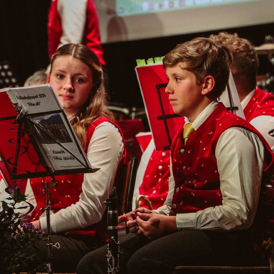 Ein junges Mädchen und ein Junge, beide in roten Westen, sitzen in einem Musikraum. Das Mädchen hat ein Notenbuch auf einem Ständer vor sich und der Junge schaut nach rechts.