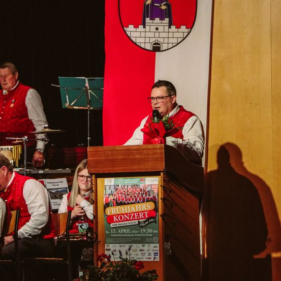 Ein Mann spricht in ein Mikrofon auf einer Bühne vor einem Banner mit einem Logo. Im Hintergrund spielen zwei Personen Instrumente. Einer sitzt, während der andere steht. Ein Plakat für ein Frühlingskonzert ist auf der Bühne sichtbar.