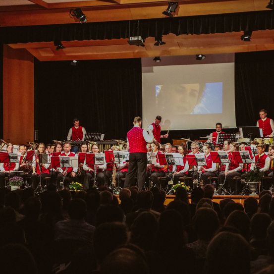 Eine Band spielt auf der Bühne bei einer Indoor-Veranstaltung, mit Musikern, die verschiedene Instrumente wie Saxophone, Flöten und Trompeten spielen. Das Publikum sitzt vor ihnen, und eine Projektionsleinwand ist an der Bühne montiert.