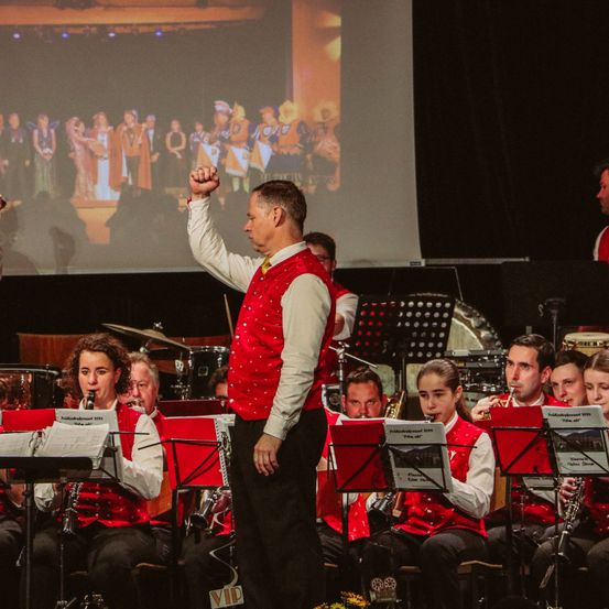 Eine Band tritt auf der Bühne auf, mit einer großen Projektionsleinwand im Hintergrund. Die Bandmitglieder tragen rote Westen, und der Mann in der Mitte macht eine Geste mit der Hand.