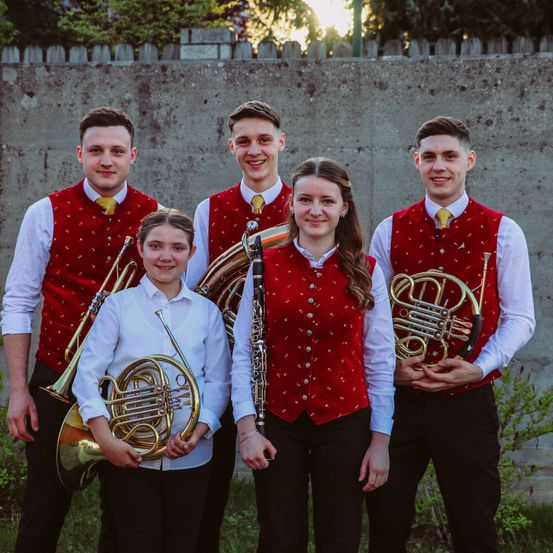 Fünf junge Musiker in roten Westen und schwarzen Hosen posieren für ein Foto. Sie halten Blechblasinstrumente.