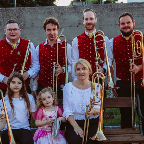 Eine Gruppe von Musikern, darunter zwei junge Mädchen, steht draußen vor einer Wand. Alle tragen rote Westen und halten Posaunen.