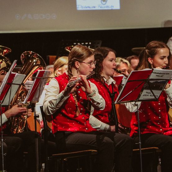 Eine Gruppe von Frauen in roten Westen und mit Brillen spielt verschiedene Instrumente, darunter Flöte, Tuba und Trompete, während einer Aufführung.