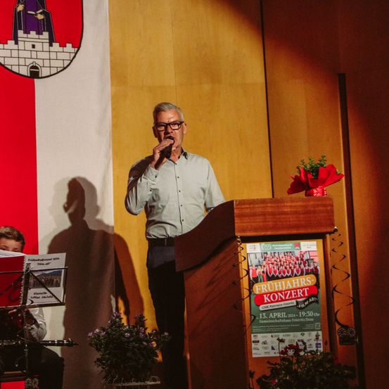 Ein Mann spricht in ein Mikrofon an einem Rednerpult mit einer Blumendekoration. Hinter ihm hängt eine rot-weiße Flagge, und ein Kind spielt im Vordergrund ein Musikinstrument.