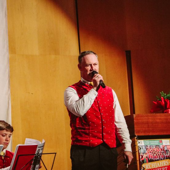 Ein Mann steht auf der Bühne und singt in ein Mikrofon. Hinter ihm sitzt ein junger Junge mit einem Notenständer.
