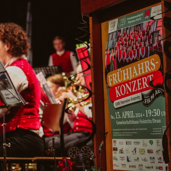 Eine Nahaufnahme eines Plakats, das ein Frühlingskonzert am 13. April 2024 um 19:30 Uhr im Gemeindehaus Feiersitz/Drau ankündigt. Eine Frau in einer roten Weste sitzt vor einem Notenständer, und eine Person im Hintergrund hält eine Trompete.