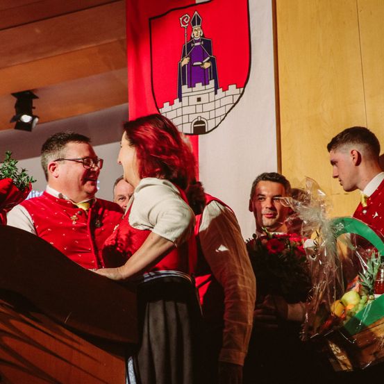 Eine Frau in einer roten Weste spricht in ein Mikrofon, während ein anderer Mann lächelt. Vor ihnen hält ein Mann einen Blumenstrauß und ein anderer Mann einen Korb mit Obst.