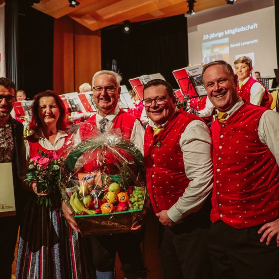 Fünf Menschen stehen lächelnd vor einer Bühne mit Musikinstrumenten. Ein Mann hält einen Obstkorb und eine Frau hält einen Blumenstrauß.