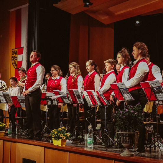 Eine Gruppe von Menschen auf der Bühne, gekleidet in rote Westen und weiße Langarmhemden, gibt ein Konzert. Sie haben Musikinstrumente vor sich.