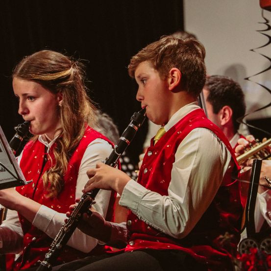 Zwei Kinder spielen während eines Konzerts Klarinette mit einem verschwommenen Publikum und einer Bühne im Hintergrund.