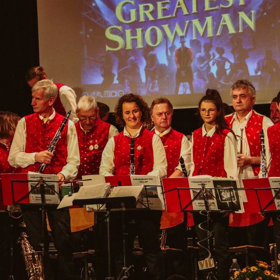 Eine Gruppe von Musikern steht auf der Bühne mit ihren Instrumenten vor einem großen Bildschirm, auf dem Greatest Showman steht.
