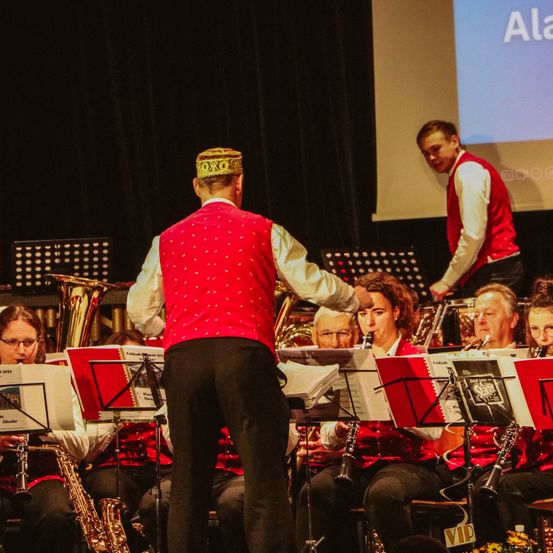 Ein Mann in einem roten Weste steht vor einer Gruppe von Musikern, die Instrumente wie Saxophone und Trompeten spielen. Ein Mann mit einer Krone steht hinter ihm.