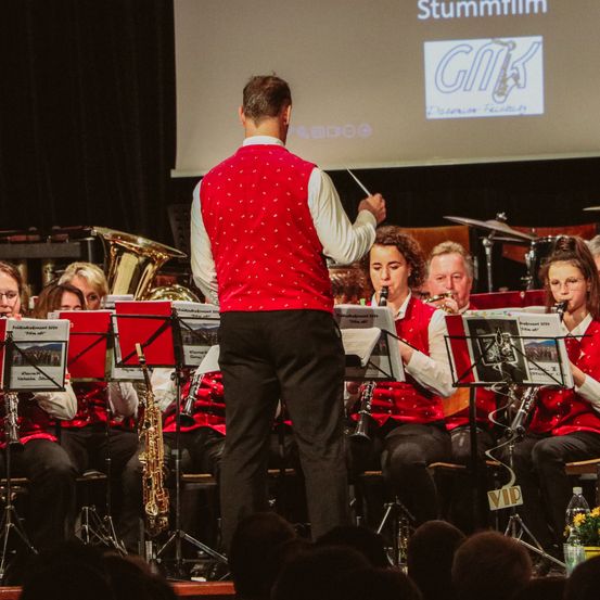 Ein Mann in einem roten Westchen dirigiert eine Band auf der Bühne mit einem Taktstock, während mehrere Musiker verschiedene Instrumente spielen, darunter Saxophone und Tuben. Das Publikum sitzt vor ihnen, und eine Projektionsleinwand zeigt das Wort 'Stummfilm'.