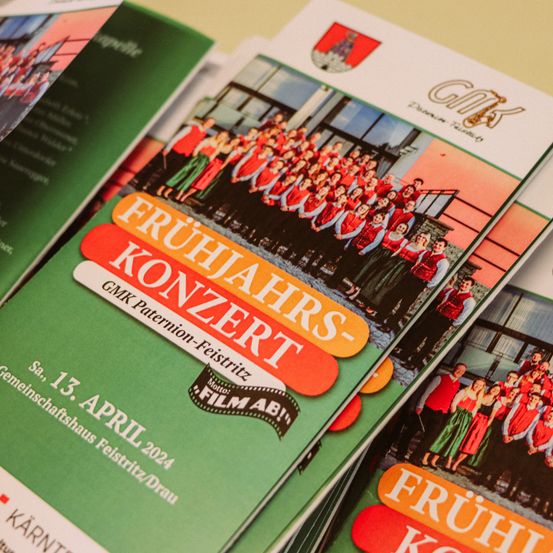 Stapel von Flyern mit Bildern von Chormitgliedern und dem Titel Fruhjahrskonzert GMK Patenion-Feistritz auf der Vorderseite.