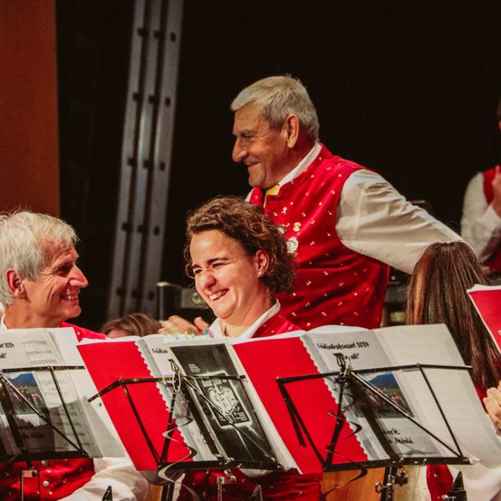 Zwei lächelnde Musiker schauen sich etwas an, während sie hinter einem Musikständer mit Notenbüchern und Notenblättern stehen.
