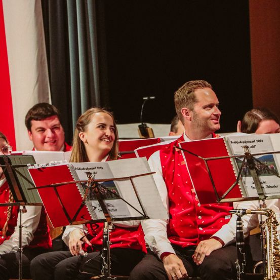 Eine Gruppe von Menschen in einer Band lächelt, und eine Person schaut in die Kamera. Die andere Person hält eine Saxophon.