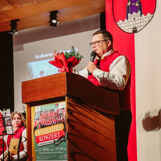 Ein Mann mit Brille und rotem Weste spricht an einem Rednerpult, während er ein Mikrofon hält. Hinter ihm steht eine Frau mit einem Buch und einem Musikinstrument. Ein Schild mit einem roten Band, ein Plakat für ein Konzert und eine Projektionsleinwand sind sichtbar.