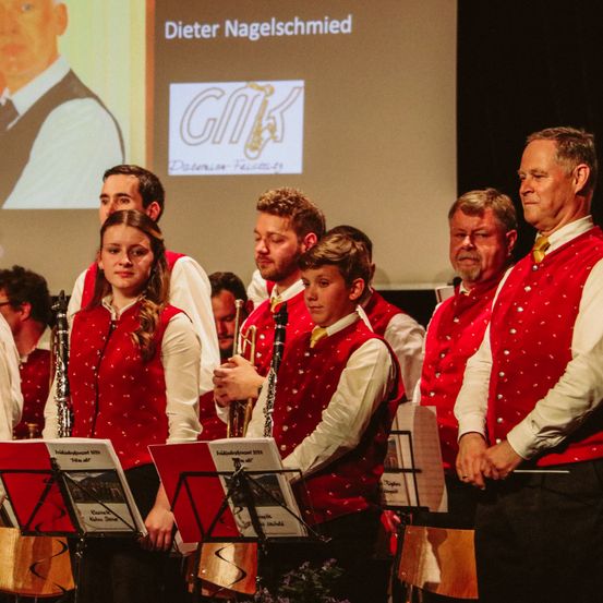 Eine Gruppe von Menschen in roten Westen mit weißen Punkten und weißen langen Ärmeln, die Musikinstrumente halten, steht vor einer Bühne mit einer Projektionsleinwand, die ein Bild eines Mannes zeigt.