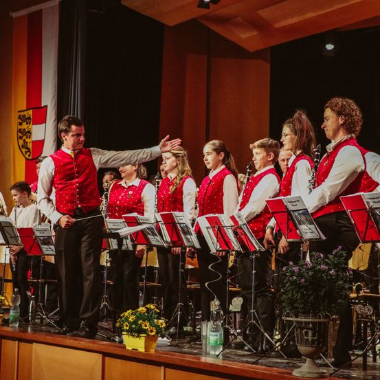 Ein männlicher Dirigent leitet eine Kinderband auf der Bühne und hält einen Taktstock in der Hand.