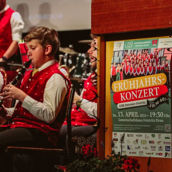 Ein junger Junge spielt Klarinette, während eine Gruppe von Musikern hinter ihm steht. An der Wand befindet sich ein Schild, das für das Frühjahrskonzert wirbt.