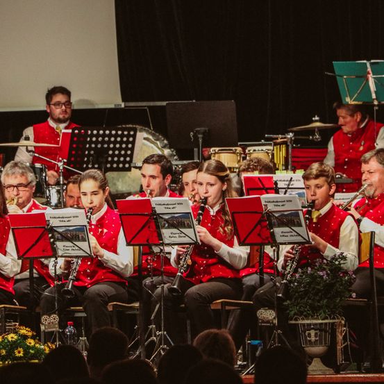 Ein Musikensemble spielt verschiedene Instrumente auf der Bühne, mit schwarzem Hintergrund und Publikum im Vordergrund.