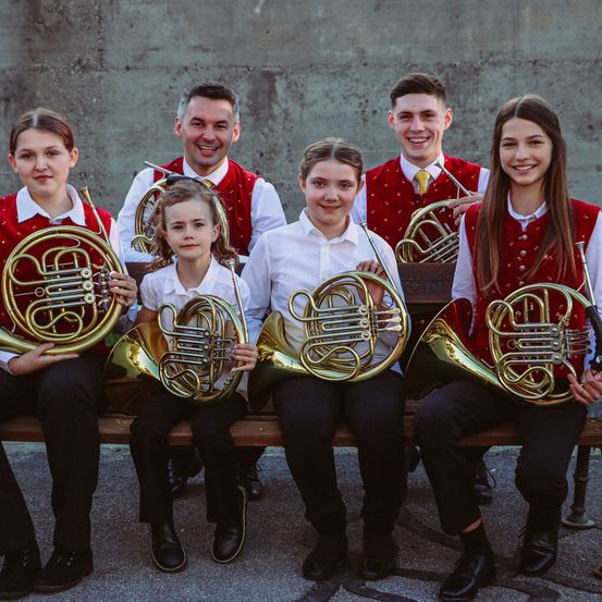 Eine Gruppe junger Musiker, die rote Westen tragen und französische Hörner halten. Zwei Frauen sitzen auf der linken Seite und drei Männer auf der rechten. Sie alle lächeln für die Kamera.