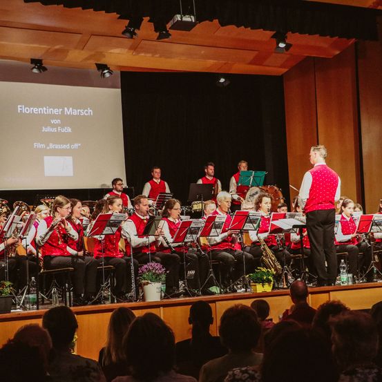 Eine Gruppe von Musikern tritt auf der Bühne auf, mit einer Projektionsleinwand dahinter, auf der 'Florentiner Marsch von Julius Fucik' steht. Der Dirigent steht vor den Musikern.