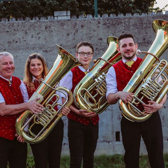 Vier Musiker in roten Westen und schwarzen Hosen halten große Messingtuben vor einer Betonwand.