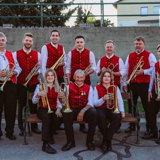 Eine Gruppe von Musikern in roten Westen und schwarzen Hosen posiert für ein Foto. Sie halten Trompeten und einige sitzen auf einer Holzbank. Sie stehen vor einer Wand und einigen Bäumen.