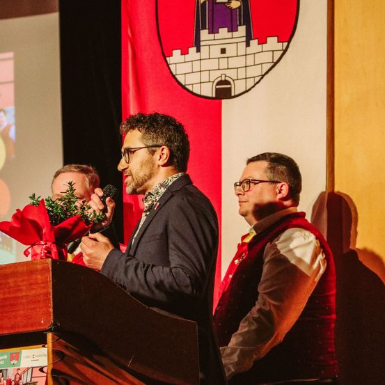 Ein Mann mit einem Mikrofon steht an einem Podium mit einem Blumenstrauß. Dahinter ist eine Flagge mit einem Burgwappen. Ein weiterer Mann in einer Weste steht zur Seite.