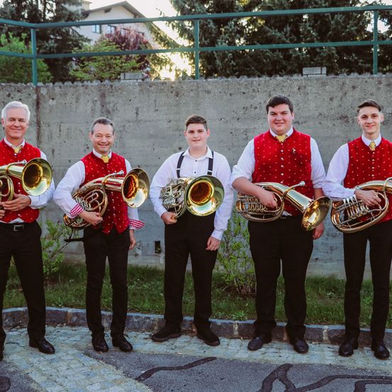 Fünf Männer stehen auf einem gepflasterten Bürgersteig in passenden roten Westen und schwarzen Hosen, halten Blechblasinstrumente und lächeln zur Kamera. Hinter ihnen befindet sich eine Betonmauer mit einem Metallzaun.