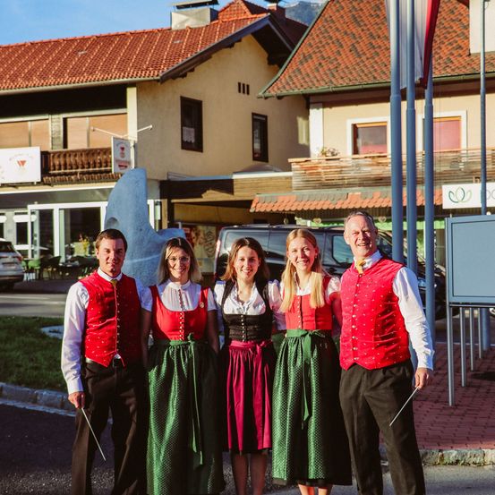 Fünf Erwachsene in traditioneller Kleidung stehen vor einem Haus. Sie lächeln und scheinen für ein Foto zu posieren. Dahinter sind eine Flagge und ein Schild zu sehen.
