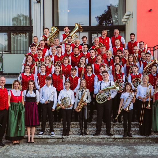 Ein Blasorchester steht vor einem Gebäude mit Glaswänden und trägt rote Westen.