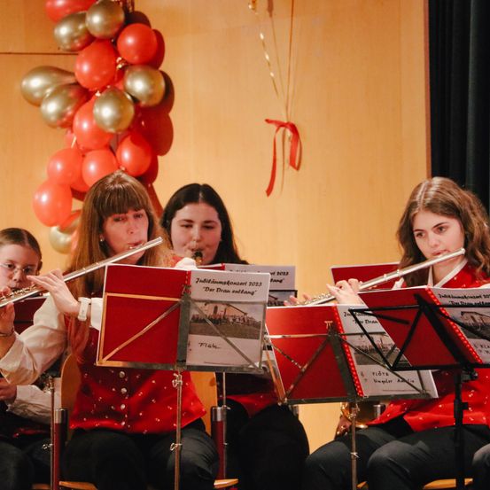 Vier Musiker sitzen in einer Reihe in roten Jacken und spielen Flöten und Klarinetten. Hinter ihnen sind ein rotes Band und eine Menge Luftballons an einer Wand befestigt.