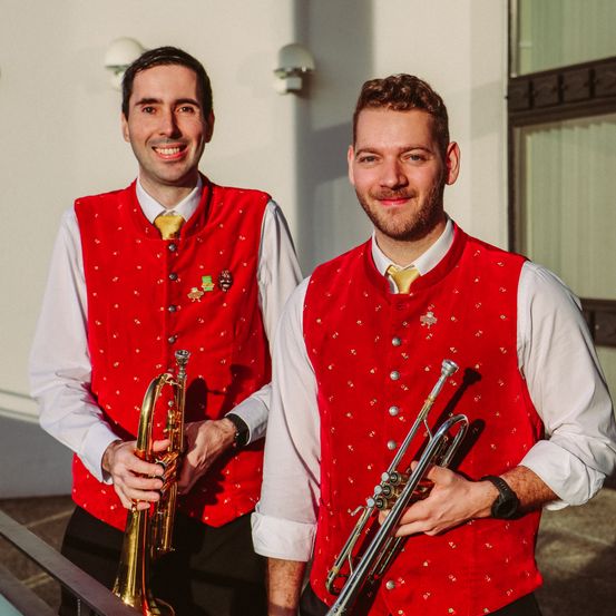 Zwei Männer in traditioneller bayerischer Kleidung, Trompeten in den Händen, stehen auf einer Treppe, lächeln und schauen in die Kamera.