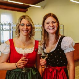 Zwei Frauen in traditioneller deutscher Kleidung lächeln und halten Weingläser, wahrscheinlich bei einer gesellschaftlichen Veranstaltung.
