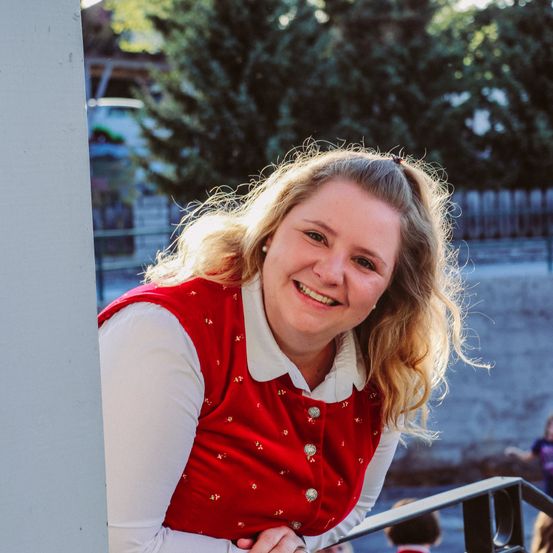 Eine Frau in einem roten Westchen steht lächelnd auf einem Balkon, mit einem Kind und einer Wand im Hintergrund.