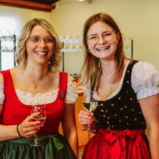 Zwei Frauen in traditioneller deutscher Kleidung, die Weinflaschen halten und lächeln, wahrscheinlich bei einer Feier oder einem festlichen Ereignis.