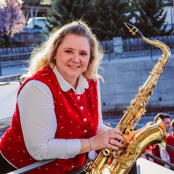 Eine Frau in einem roten Weste mit weißem Rand und einem weißen langärmeligen Hemd steht auf einem Balkon, lächelt und hält eine goldene Saxophon. Hinter ihr ein verschwommener Hintergrund von Bäumen, einer Betonmauer und einem Gebäude.