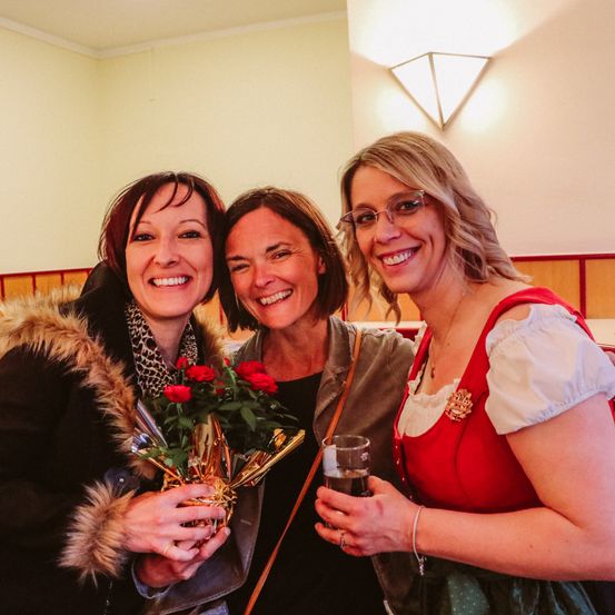 Drei Frauen stehen zusammen, zwei von ihnen lächeln, eine mit einem Strauß roter Rosen, eine andere mit einem Glas Bier und eine dritte in einem roten Kleid, wahrscheinlich bei einem festlichen Ereignis.