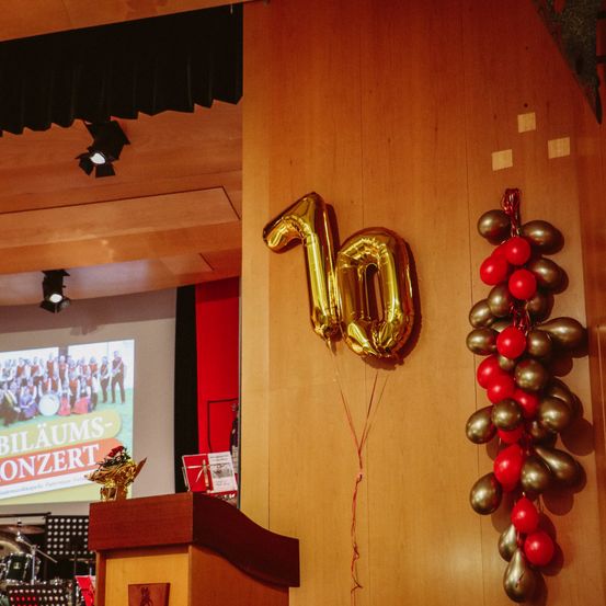 Ein goldener Luftballon mit der Nummer 10 hängt an einem braunen Pfeiler, rechts befindet sich eine dekorative Anordnung aus roten und goldenen Luftballons.