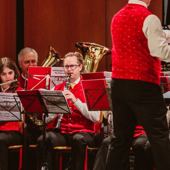 Eine Gruppe von Musikern tritt in einem Konzertsaal auf. Der Mann in der Mitte spielt Klarinette. Die beiden Personen auf der linken Seite spielen Trompete und Posaune. Der Dirigent geht hinter ihnen.