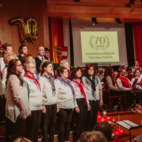 Ein Chor in weißen und schwarzen Kleidern singt vor einem Publikum. Dahinter zeigt eine Leinwand '70 Jahre Gemeindemusikkapelle'. Musikinstrumente stehen auf der Bühne.