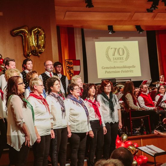 Eine Gruppe von Frauen in weißen Shirts und roten Schals singt auf der Bühne mit einem Chor hinter ihnen. Sie stehen vor einer Projektionsleinwand, auf der '70 Jahre 1953-2023' und 'Gemeindemusikapelle Paternio-Feistritz' steht.
